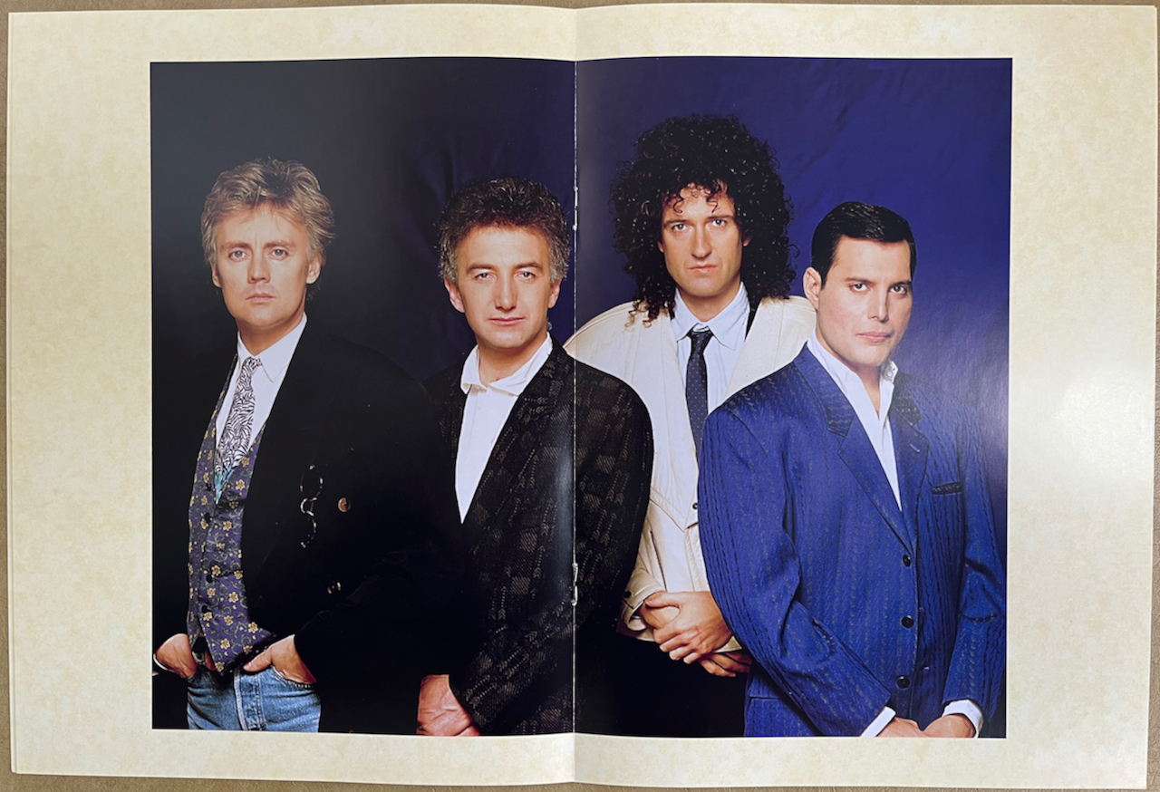 A double-page spread from the biography booklet in Queen's box set for The Miracle, showing a large colour photo of the band posing together. Freddie is in a blue suit with a white shirt. Brian is wearing a cream jacket over a white shirt and a navy tie, John is wearing a black jacket over a white shirt, and Roger is wearing a black jacket over a patterned waistcoat and tie and a white shirt.