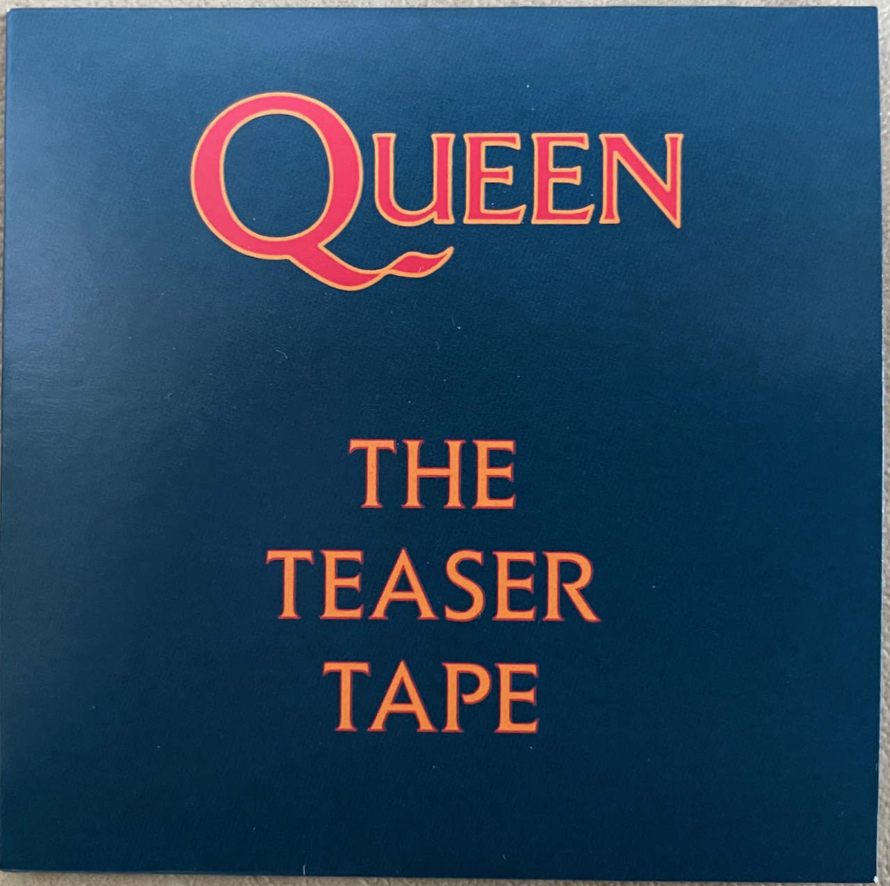 Front cover of the small cardboard sleeve for the mini CD teaser tape of The Miracle. Against a dark blue background, the Queen logo is in red text with thin yellow edging, while the words The Teaser Tape are in yellow letters with thin red edging.