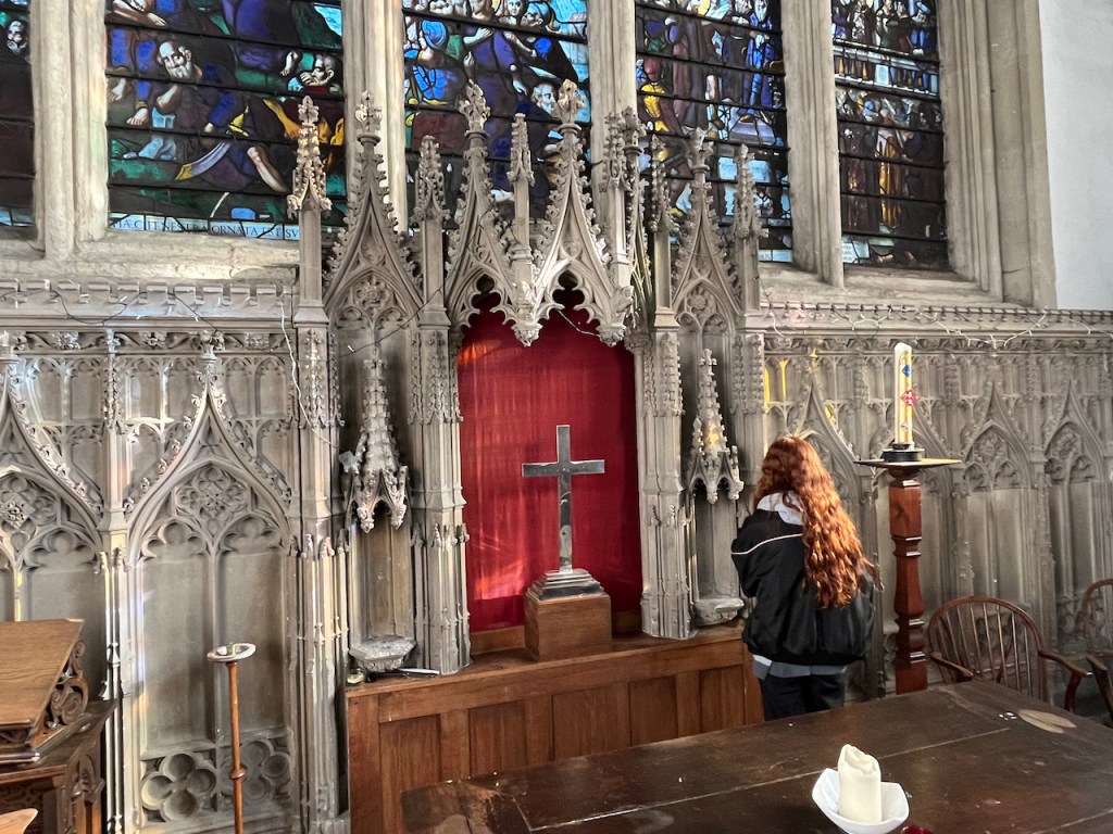 Impressively detailed architecture below the stained glass window in Wadham College chapel, including heavily detailed triangular spikes surrounding an opening containing a metal cross.