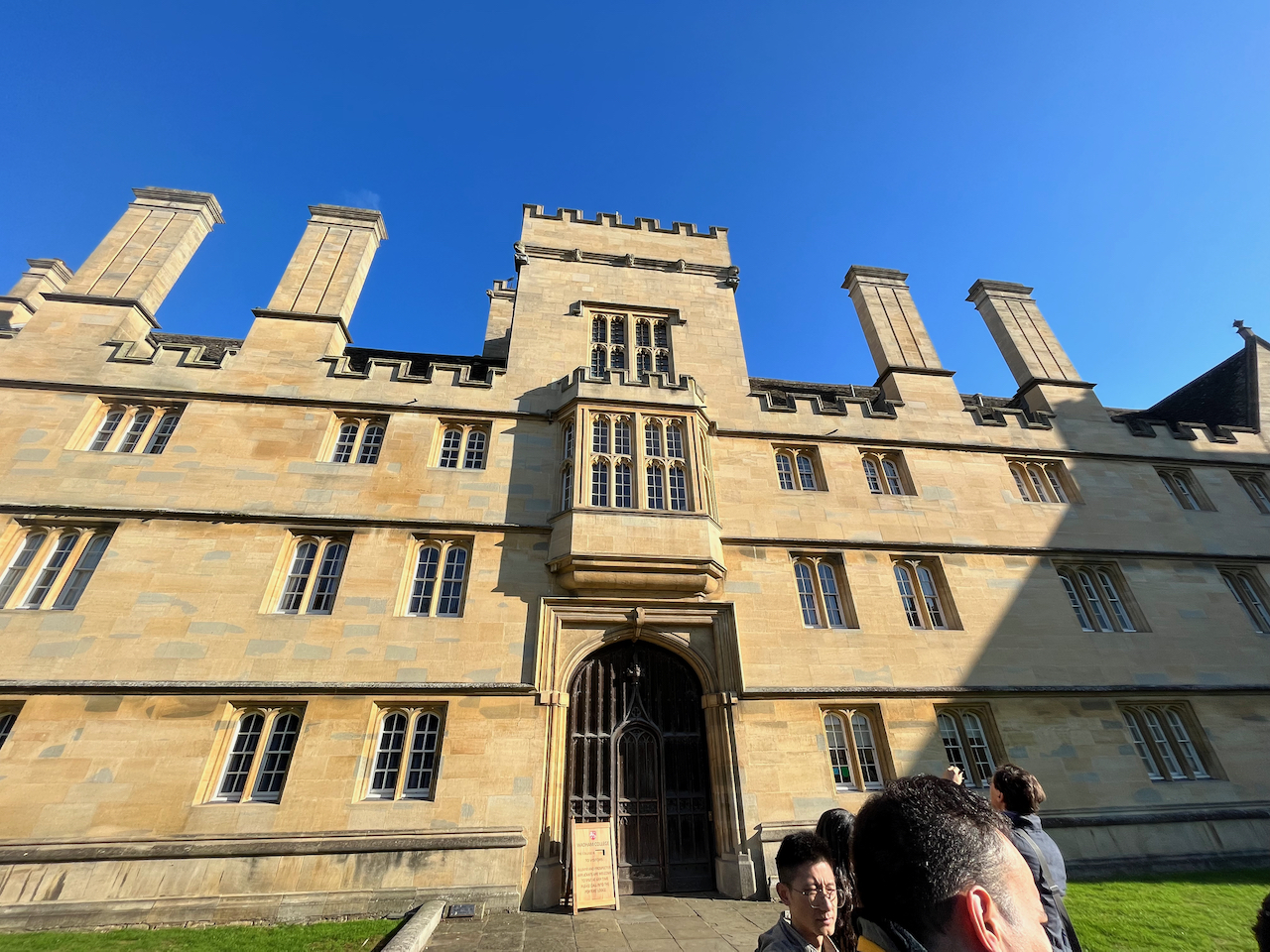 The exterior of Wadham College, a mainly 3 storey building, but with some chimney-like protuberances at regular intervals across the top, and a 2 storey tower above the tall entrance archway.