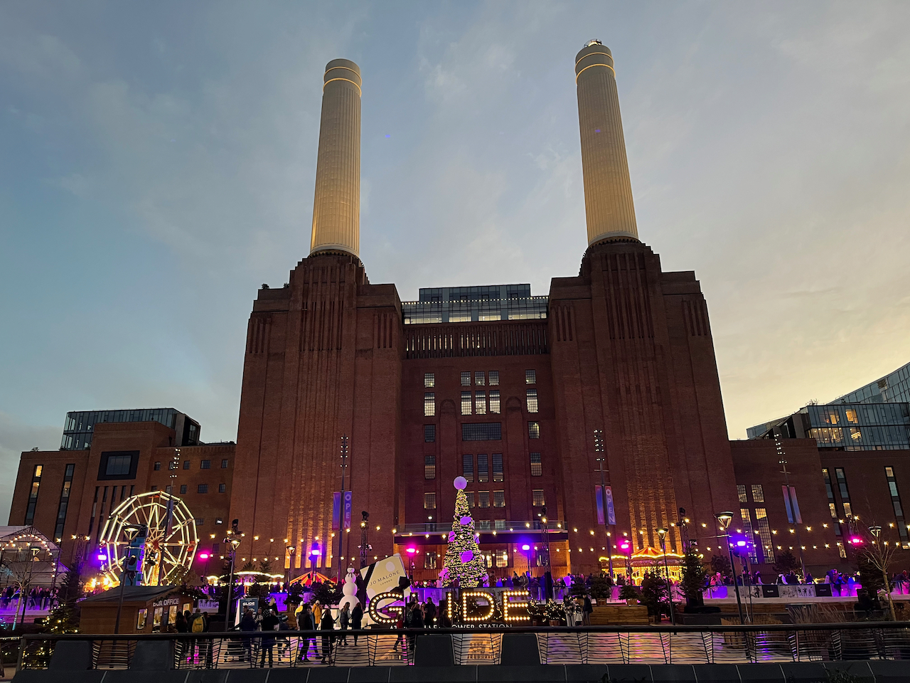 Battersea Power Station, a huge building with 2 enormous chimney towers, one on each side. On the ground in front of it are Christmas lights, a tree, an ice rink and a fairground, all looking bright and colourful.