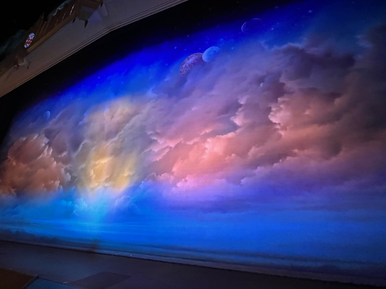 The stage for The Book Of Mormon, with the curtain showing a colourful scene of clouds with sunlight poking through, above which the moon and some stars are visible.