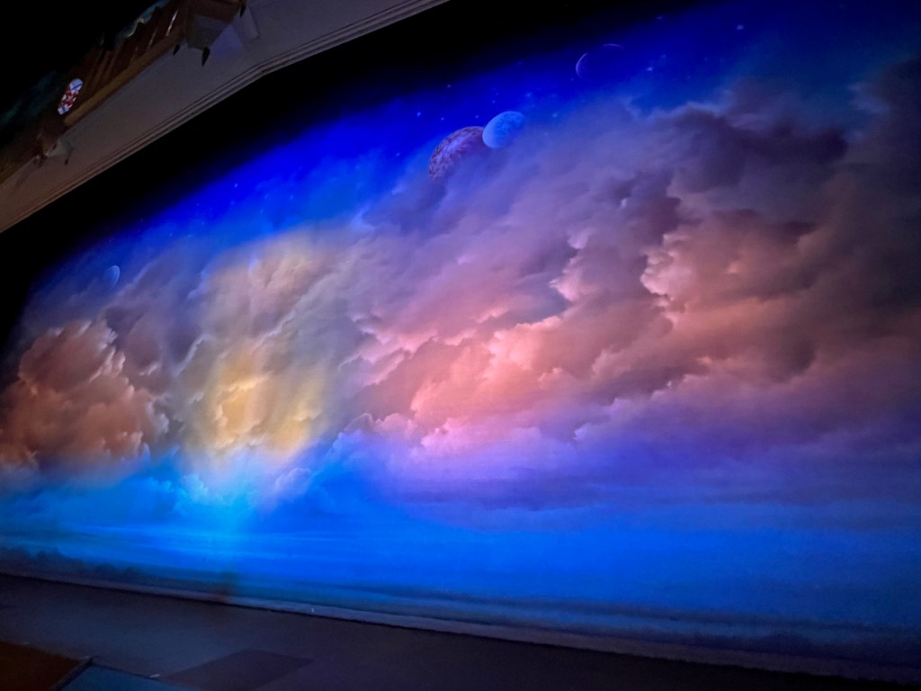 The stage for The Book Of Mormon, with the curtain showing a colourful scene of clouds with sunlight poking through, above which the moon and some stars are visible.