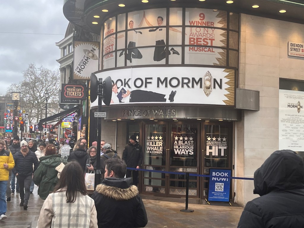 The entrance to the Prince Of Wales Theatre, with a banner for the Book Of Mormon above it. The second O in the word Mormon is replaced by a doorbell, and beneath the wording a guy wearing a white shirt and black trousers is relaxing, stretched out on his side, as he reads a copy of the Bible. Above that banner is a larger poster covering a window, with 2 guys in white shirts and black trousers happily jumping and high-fiving each other, while text either side of them says it's won 4 Olivier Awards and 9 Tony Awards.