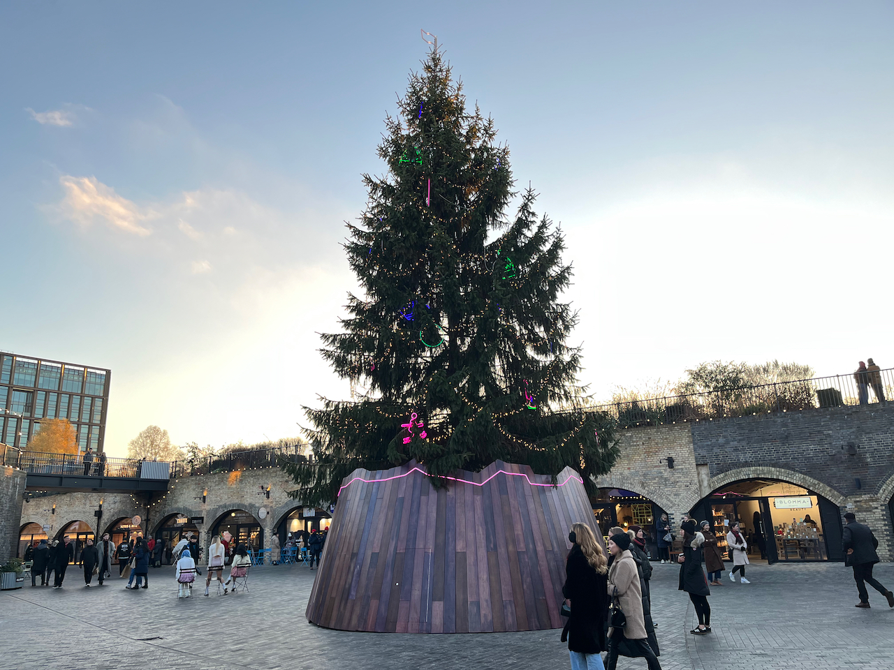 A tall green Christmas tree covered in strings of lights and colourful decorations on a large round wooden base, outdoors in Coal Drops Yard in front of a line of stone archways.