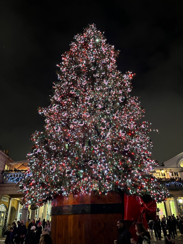 The huge Christmas tree covered in twinkling lights and reflective silver baubles in Covent Garden, towering over the buildings around it.
