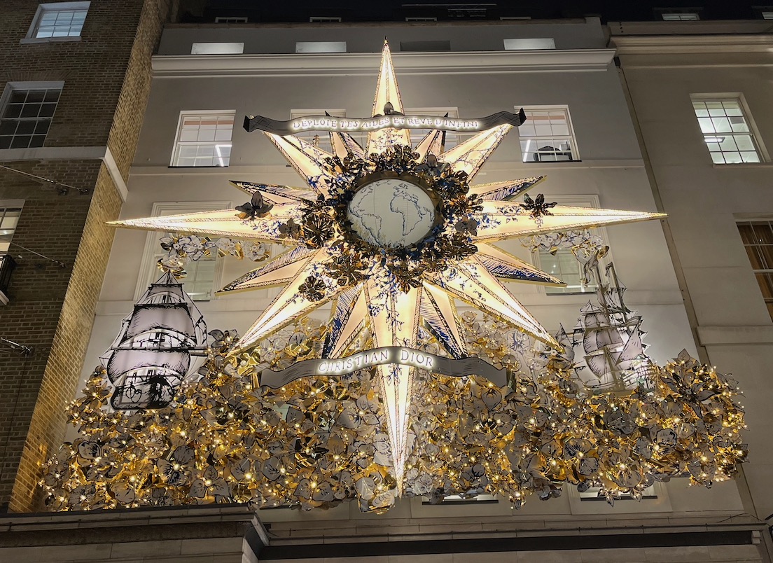 A brightly lit 16-point star. The top, bottom, left and right points of the star are the largest and longest, while the others in between are shorter. In the centre of the star, surrounded by a decorative wreath, is a globe with a map of the Earth on it. Latin text across the top of the star reads Déploie tes ailes et rêve d’infini, meaning Spread your wings and dream of infinity.