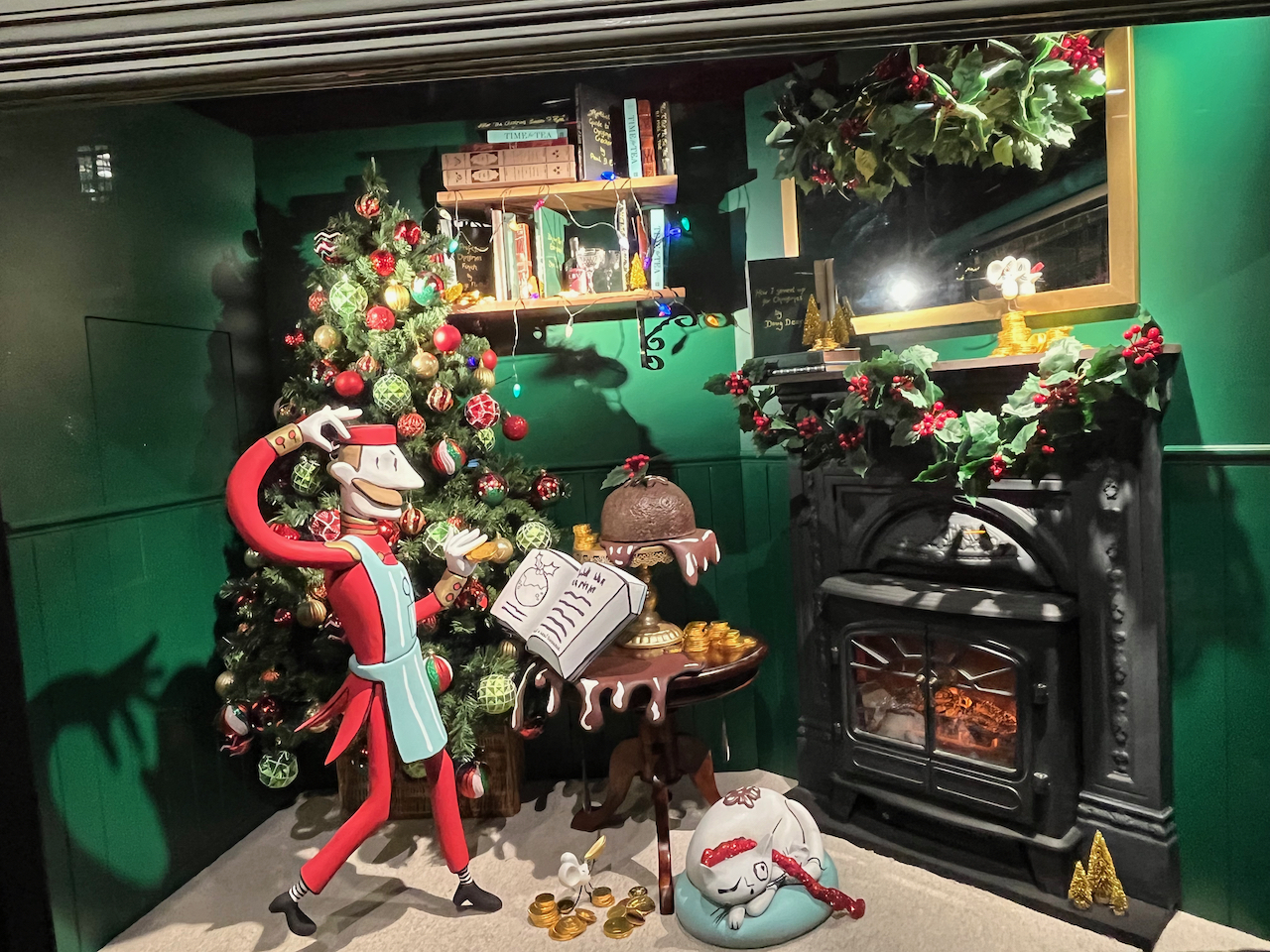 Fortnum and Mason window display of a chef, who has a moustache and is dressed in red with a white apron, standing in front of a Christmas tree and reading a recipe for a Christmas pudding. A pudding is sitting on the table in front of him, next to a stove. A white cat is curled up on a cushion in front of the stove, with one eye open as it looks around.