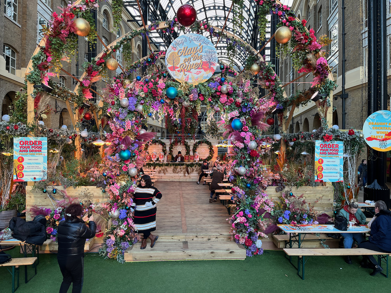 A bar in Hay's Galleria called Hay's On The River. The archway leading into it is covered in baubles and decorations in pink, purple and blue colours, with further wooden arches across the space behind it that are also covered in decorations. The wooden bar at the back of the space has decorations along the front of it, and green wreaths on the wall behind the bartender.