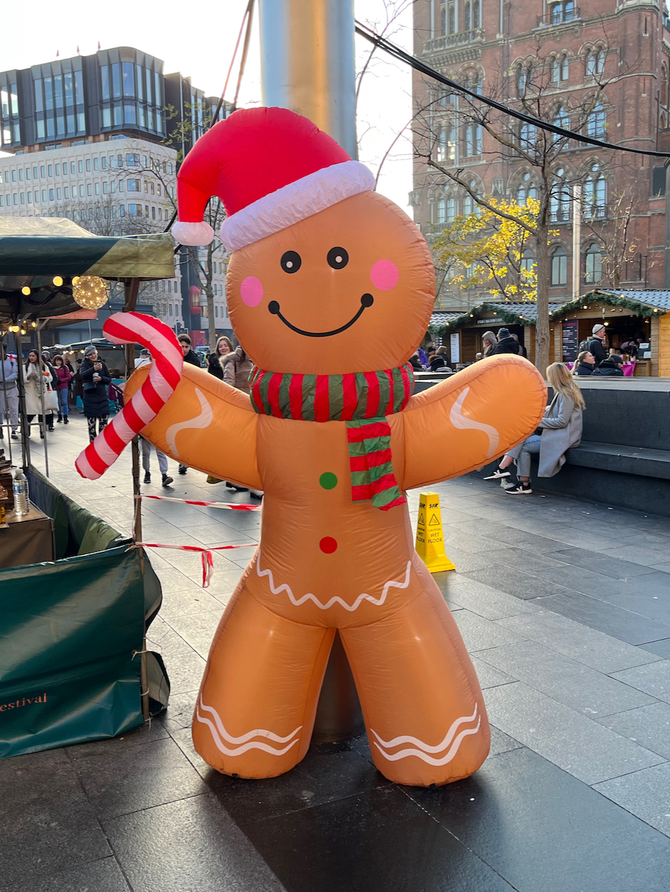 A large inflatable model of a smiling, rosy-cheeked gingerbread man, wearing a green and red striped scarf and a red and white Santa hat, and holding a red and white striped candy cane in its right hand, in the Christmas market by Kings Cross Station.