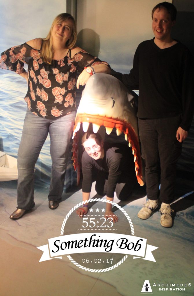 Glen and Louisa smiling and posing either side of a large model of a shark's open mouth, while Jody smiles while on hands and knees inside the mouth. Their escape room team name, Something Bob, is shown at the bottom of the photo, with their completion time of 55 minutes and 23 seconds.