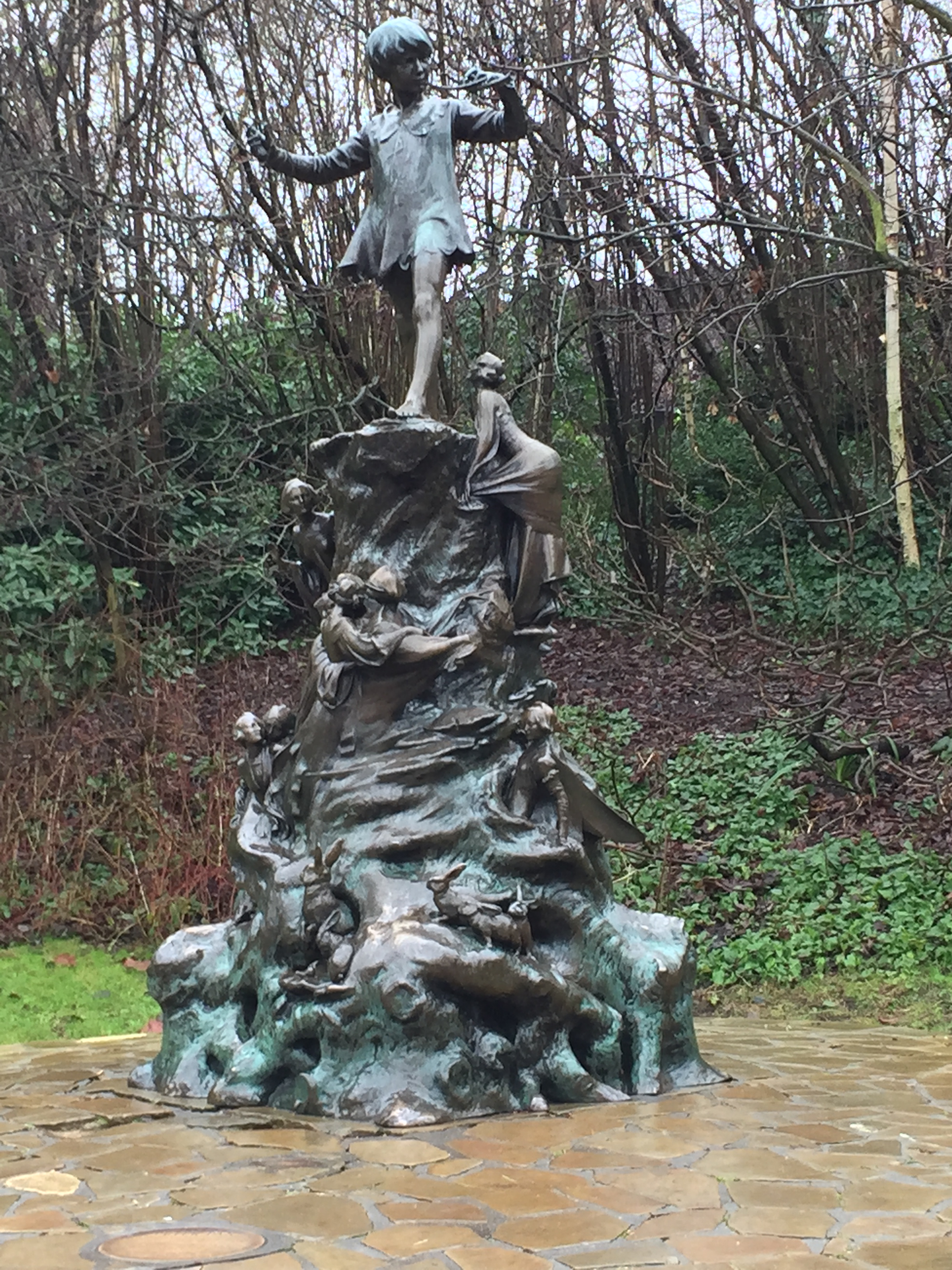 A bronze statue of Peter Pan standing on a tall rock, surrounded by fairies, squirrels, rabbits and mice.