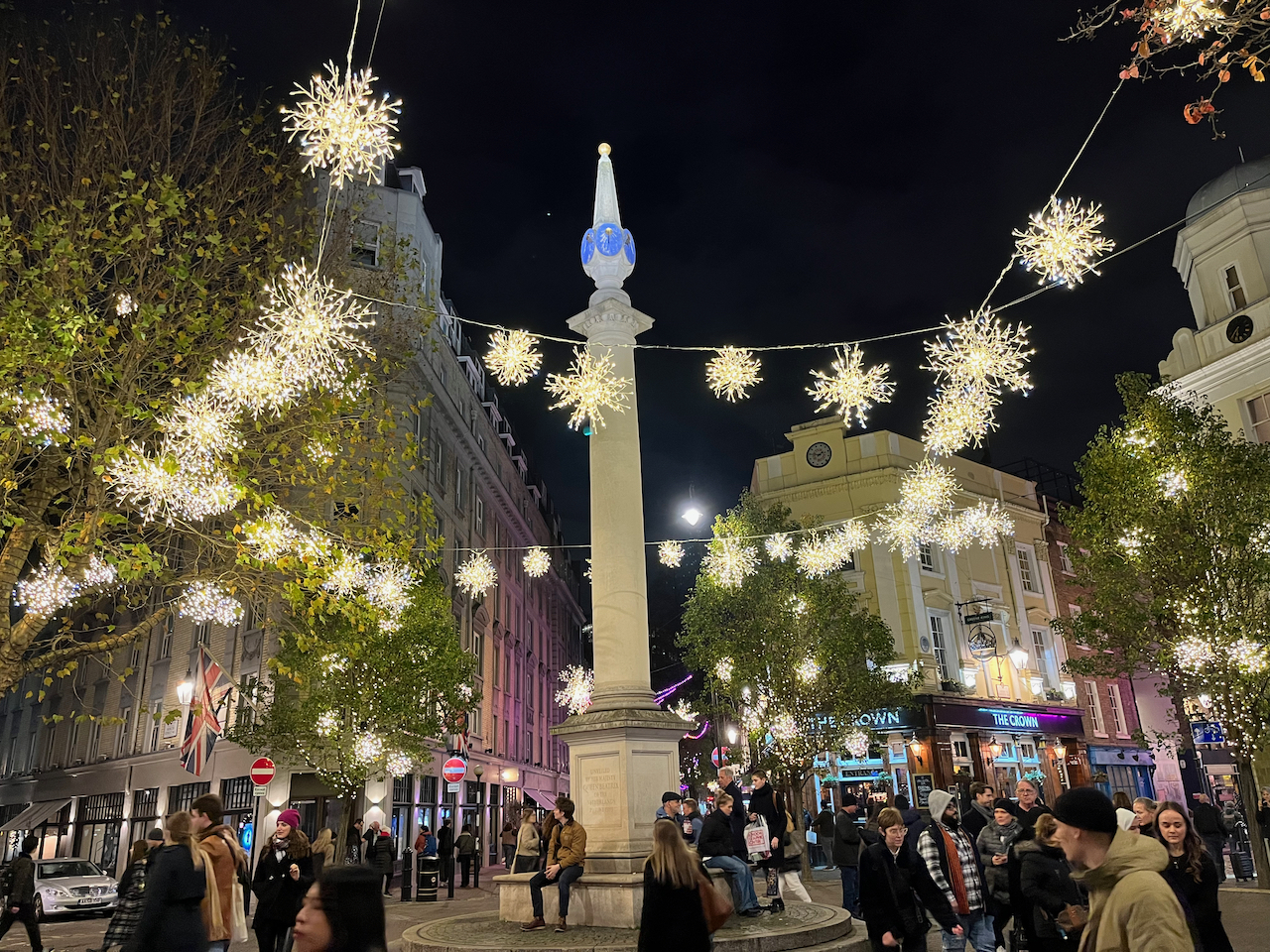 The tall column with a rounded spire in the centre of the Seven Dials shopping district. Surrounding it are cables holding lots of brightly coloured lights that look like exploding snowflakes.