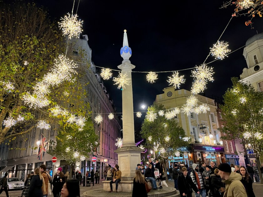 The tall column with a rounded spire in the centre of the Seven Dials shopping district. Surrounding it are cables holding lots of brightly coloured lights that look like exploding snowflakes.