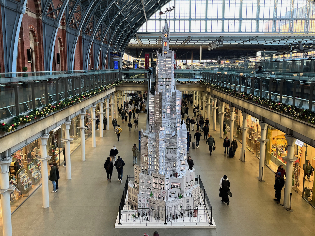 A 33-foot tall tall tree-like structure in St Pancras International station, made entirely of 2D representations of London buildings drawn in black on white card, including houses, churches, shops, ferris wheels, bridges, theatres, tower blocks, monuments, etc. Lighting inside the tree is visible through the many little windows in the buildings. There are also lots of little details including silhouettes of people on the streets and through the windows, phone boxes, drawings of landscapes, and green Christmas trees and decorations. At the very top, a silhouette of Santa being pulled by his reindeer flies over the top of a Big Ben style clock tower.
