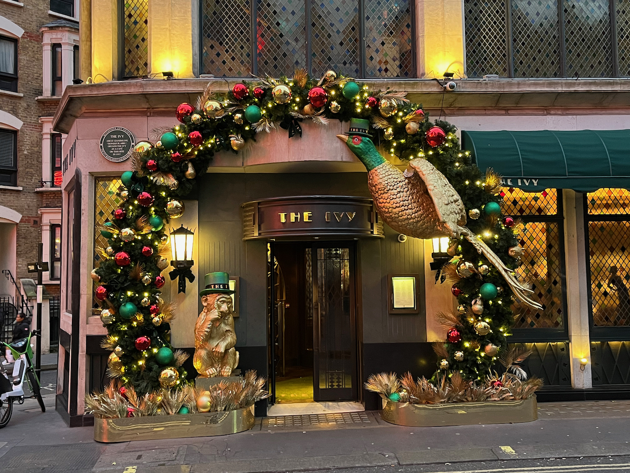 The entrance to The Ivy, surrounded by an archway of greenery heavily decorated with red and gold baubles and festive lights. Hanging under the right side of the arch is a large gold pheasant with a long tail, wearing a green top hat on its head. On the ground by the left side of the archway is a gold statue of a monkey in a thoughtful pose, also wearing a top hat.