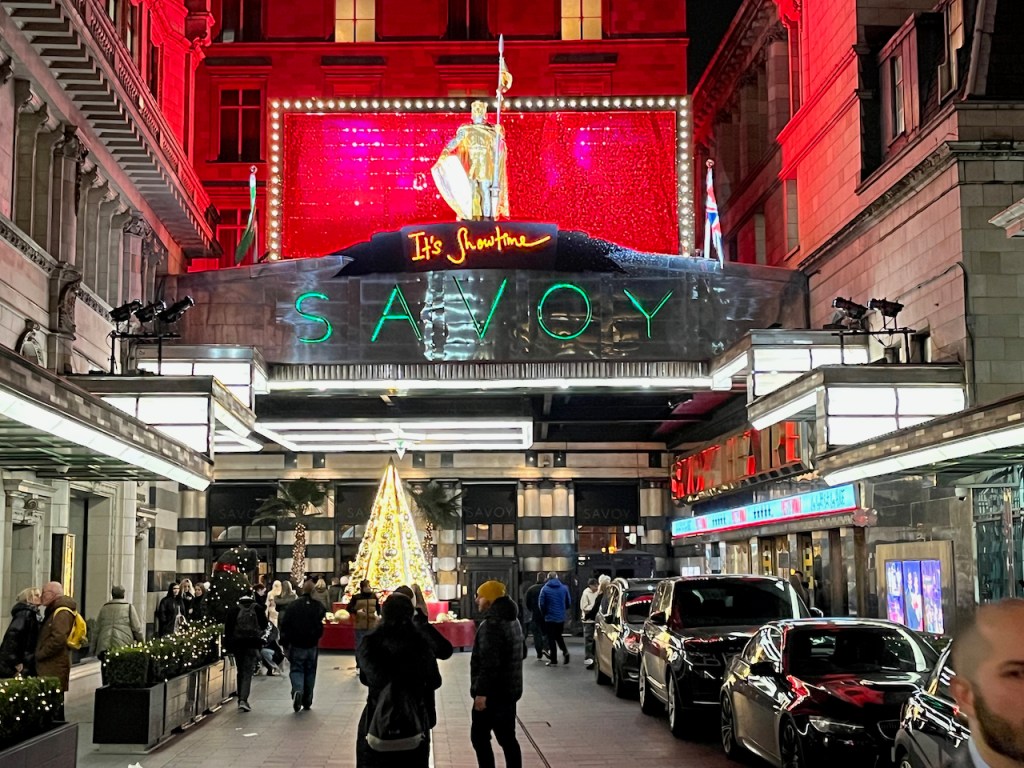 The entrance road leading to the Savoy hotel, with its upper floors lit up in red. A figure holding a shield and a flag stands above the entrance canopy in the distance, with the words It's Showtime just below the figure in bight handwritten style letters. The word Savoy is in big gold capital letters across the front of the canopy.