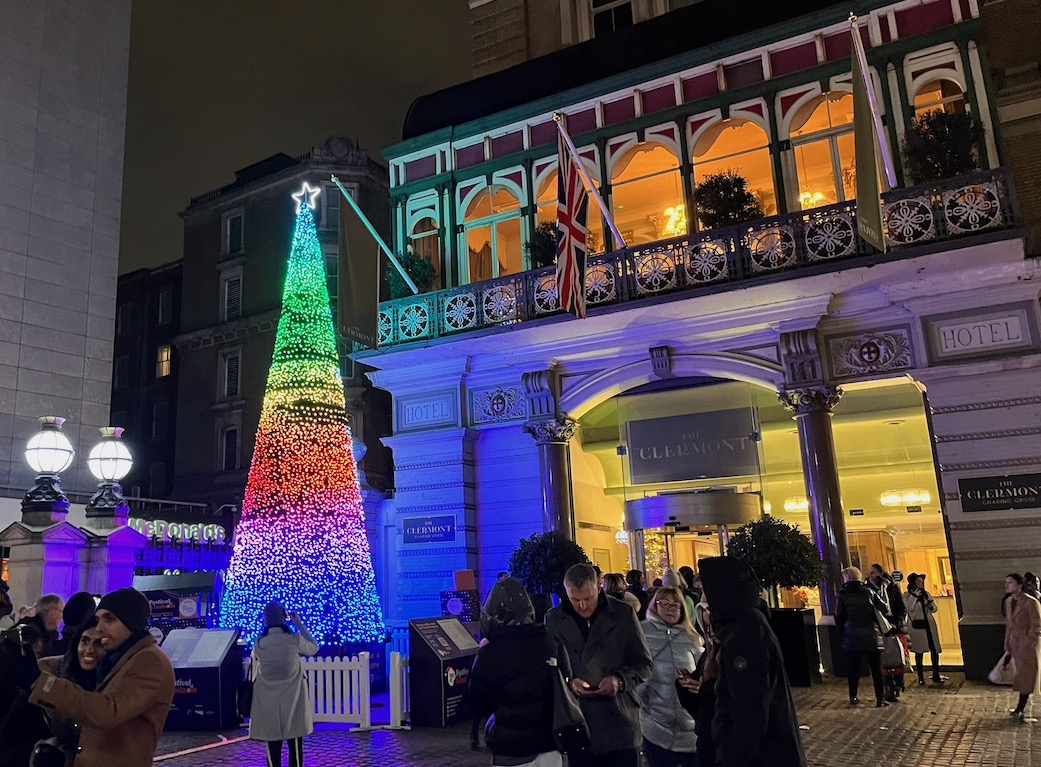 The Tree Of Kindness, a very tall tree made completely of lights that change colour in a variety of patterns, with a white star on top. Here the tree is displaying a rainbow of colours in horizontal bands that fade into each other, from top to bottom in violet, purple, blue, green, yellow and red.