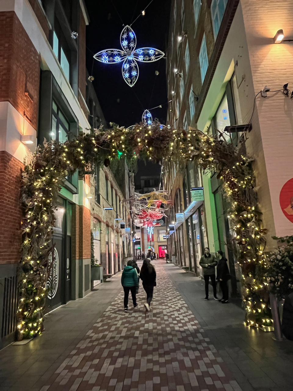 An archway covered in greenery and festive lights, above which is a lit Christmas decoration shaped like a 4-leaf clover. The archway leads to a pedestrianised shopping street which has further colourful decorations overhead in the distance.