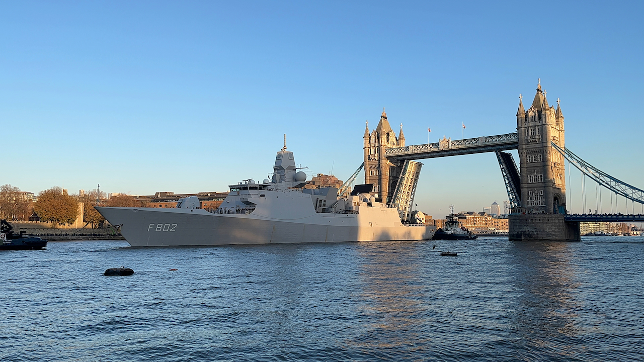 A huge grey warship, with the code F802 in white on the side and people visible on deck, passing under Tower Bridge, which has the 2 sections of the road lifted open in the centre, all under a clear blue sky.