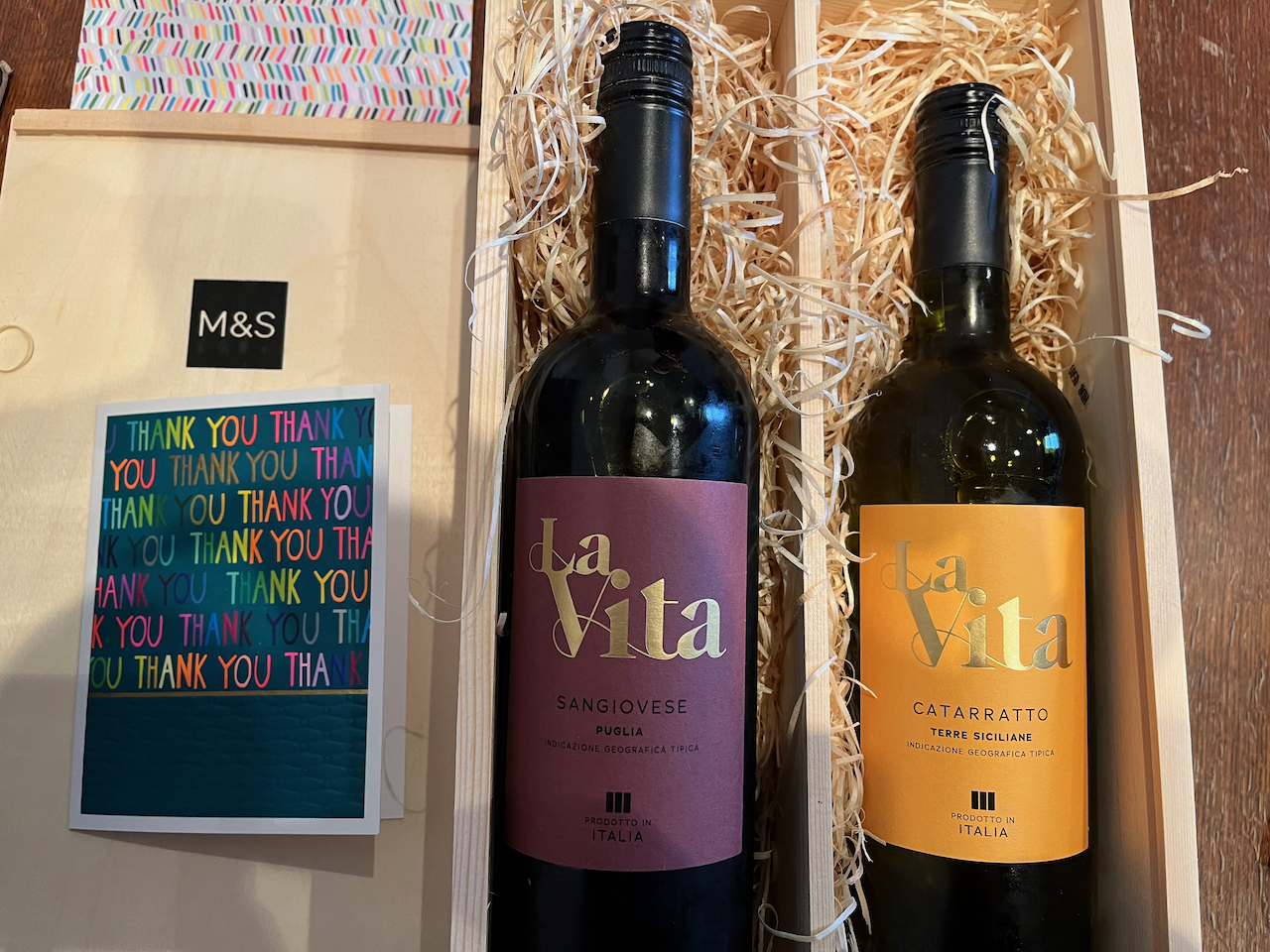 2 bottles of La Vita wine, one red and one white, in a wooden box from M&S, alongside a card that has the words Thank You written many times on the front in different coloured letters.