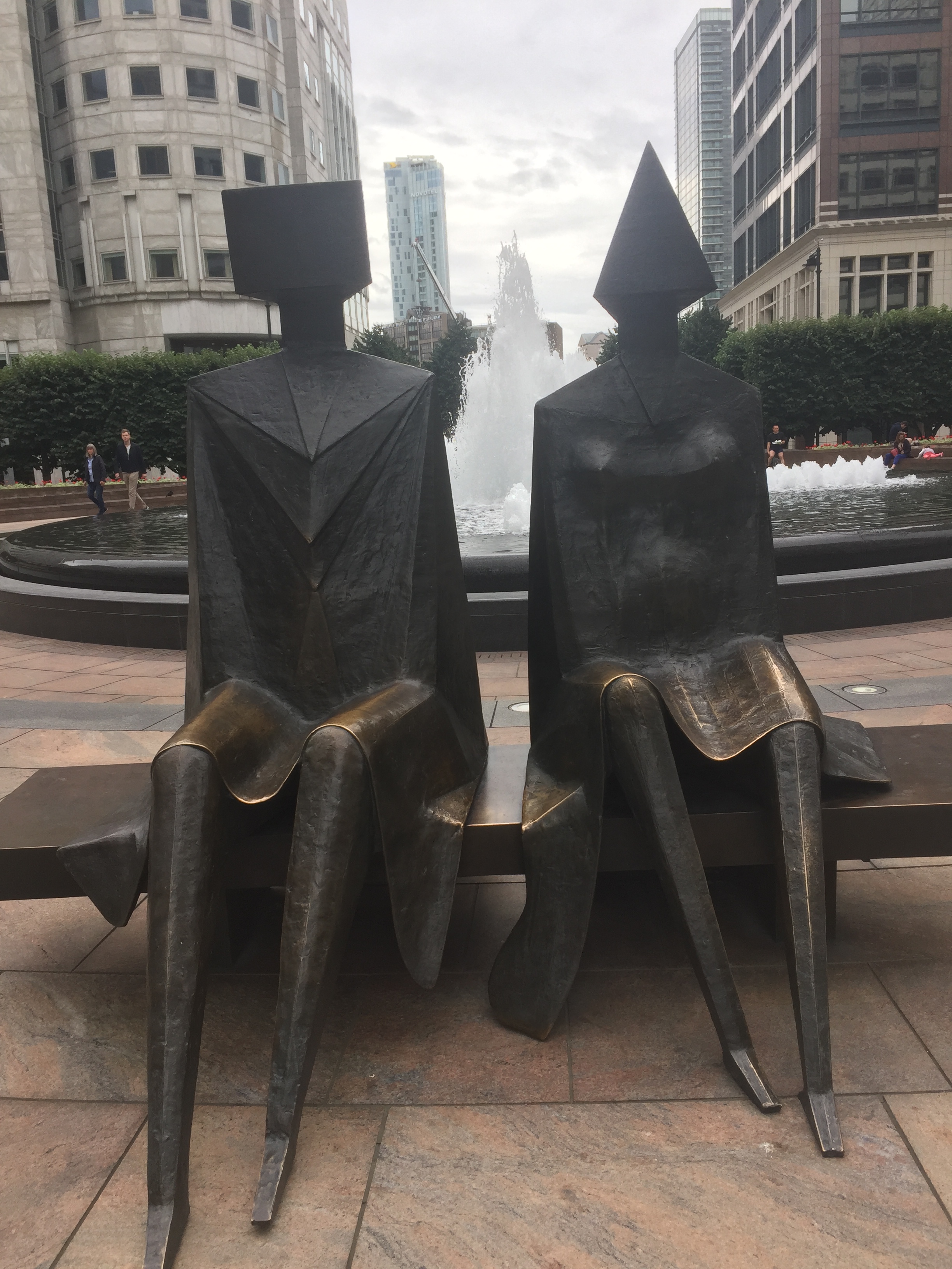 Artistic bronze sculptures of a couple sitting on a bench in Canary Wharf. The figures aren't designed to be lifelike, as they have rectangular and triangular heads respectively, very straight shoulders, no arms, thin legs, and both appear to be wearing skirts. A large fountain, contained in a large round pool, can be seen spraying behind them, through the gap in between them.