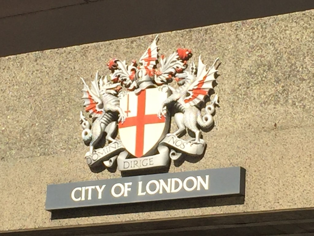 A sculpture of he City Of London coat of arms on a wall. The centrepiece is a shield with a white background and a red cross, with an image of a sword in the top left quadrant formed by the cross. A dragon stands on each side of the shield, facing inwards towards it. A knight's helmet sits on top of the shielded, with the red and white flag sticking out of it. Below the shield is the motto Domine dirige nos, meaning Lord guide us.