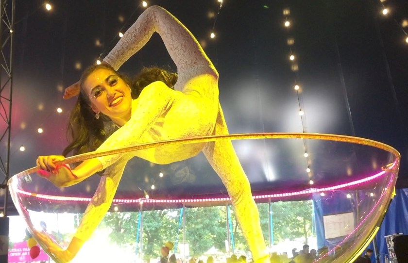 A female contortionist in large model of a martini glass, smiling as she holds on to the rim and puts one leg over her head.