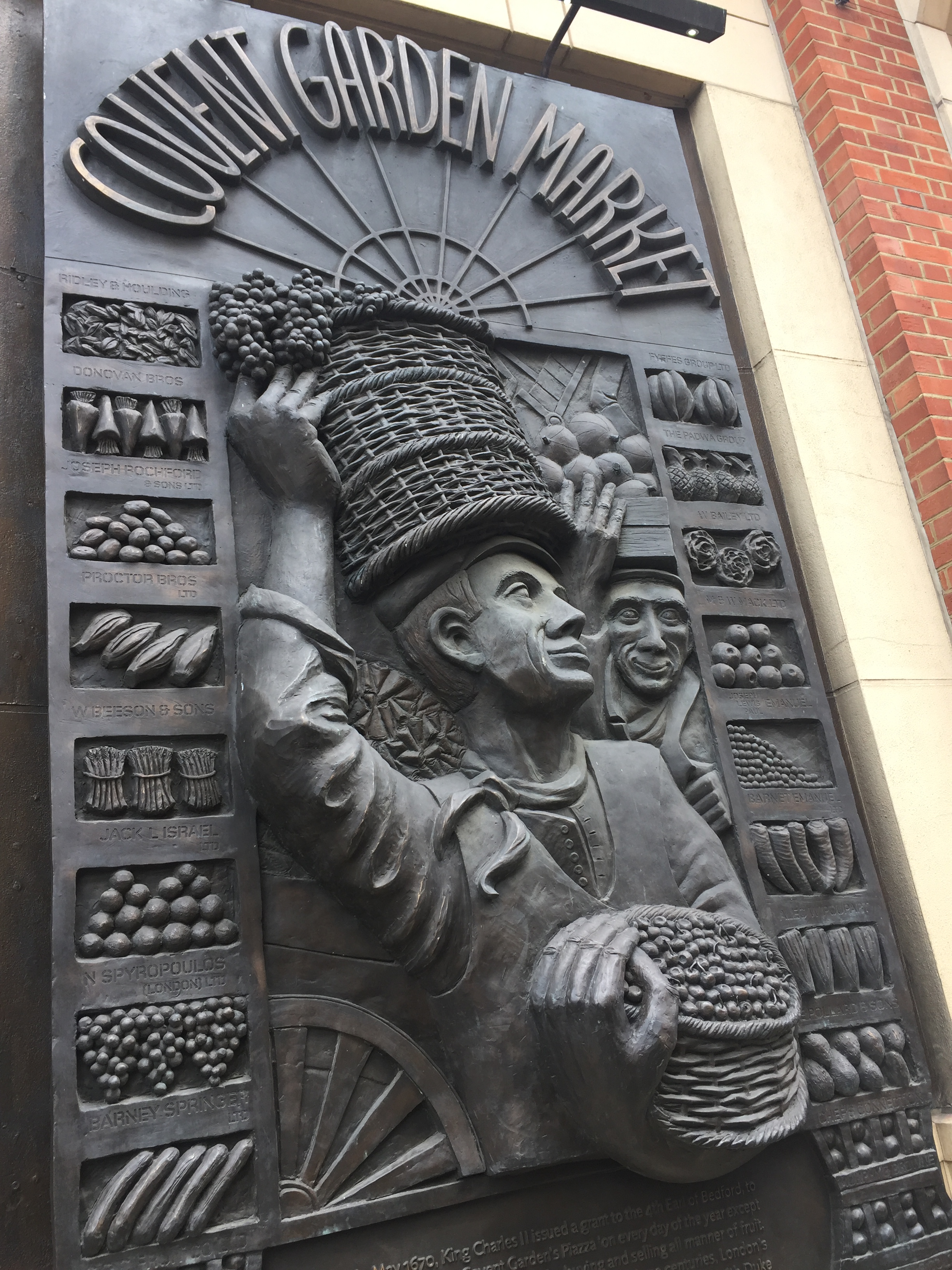 A large sculpture in the form of a sign for Covent Garden Market, showing a man holding a large basket of fruit on his head with his right arm, while his left arm is wrapped underneath another basket of fruits.
