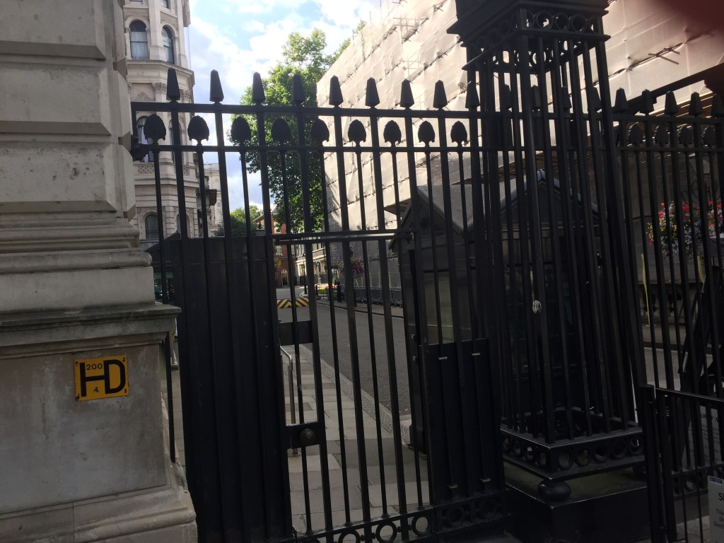 Looking through the tall black gates of Downing Street, with the area outside 10 Downing Street just visible in the distance beyond a yellow and black barrier in the road.