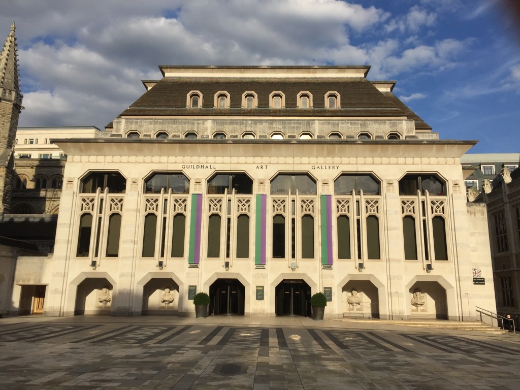 The large Guildhall Art Gallery building, with arches at ground level that are formed of straight lines rather than curves, with diagonals linking the vertical sides to the horizontal top section. Above each of those arches are 2 tall but narrow vertical windows side by side, with a slightly larger window across their width above.