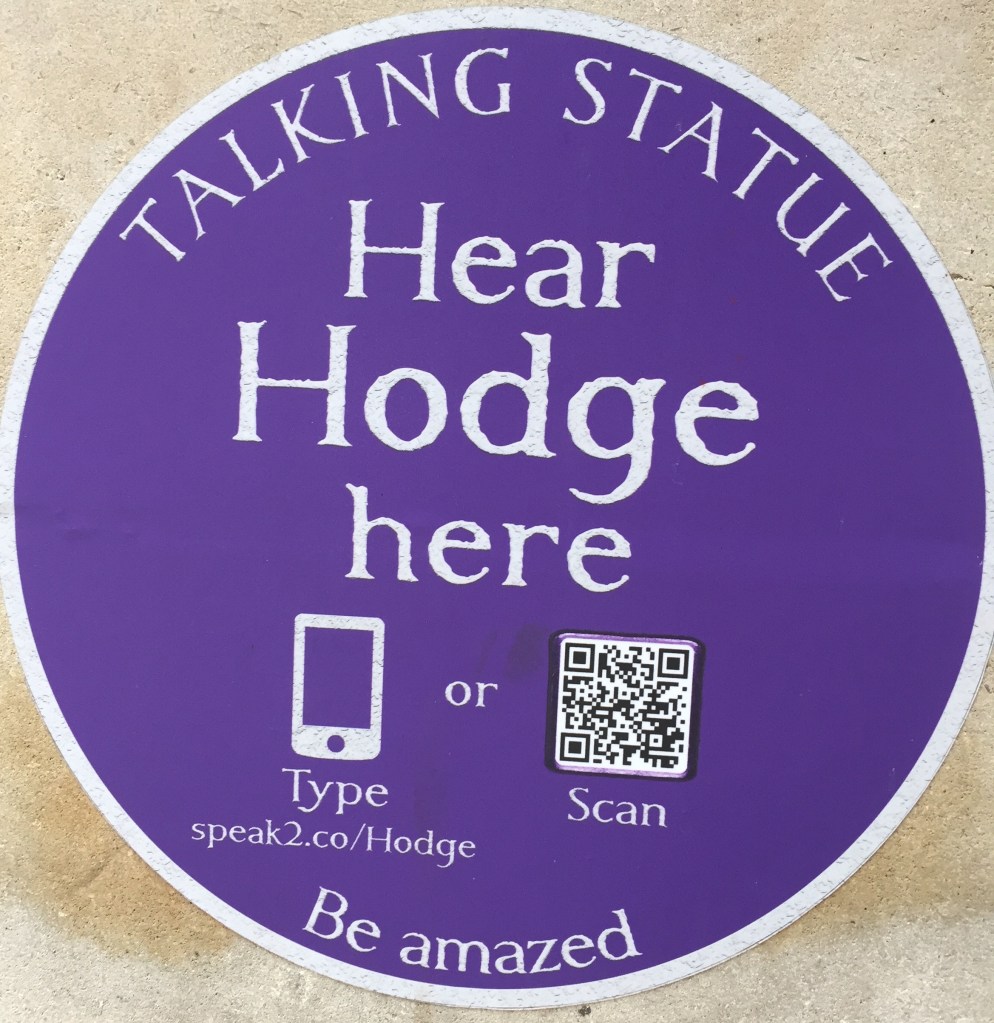 A round blue plaque with white text that says Talking Statues, hear Hodge here, be amazed. There is a QR code that scan be scanned, or a short web address that can be typed in.
