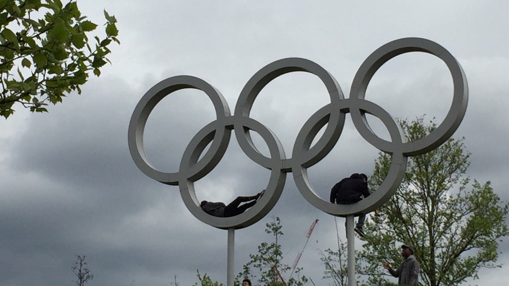 Large grey metal sculpture of the Olympic rings. The 2 bottom rings, supported on tall poles, each have a person laying inside them.