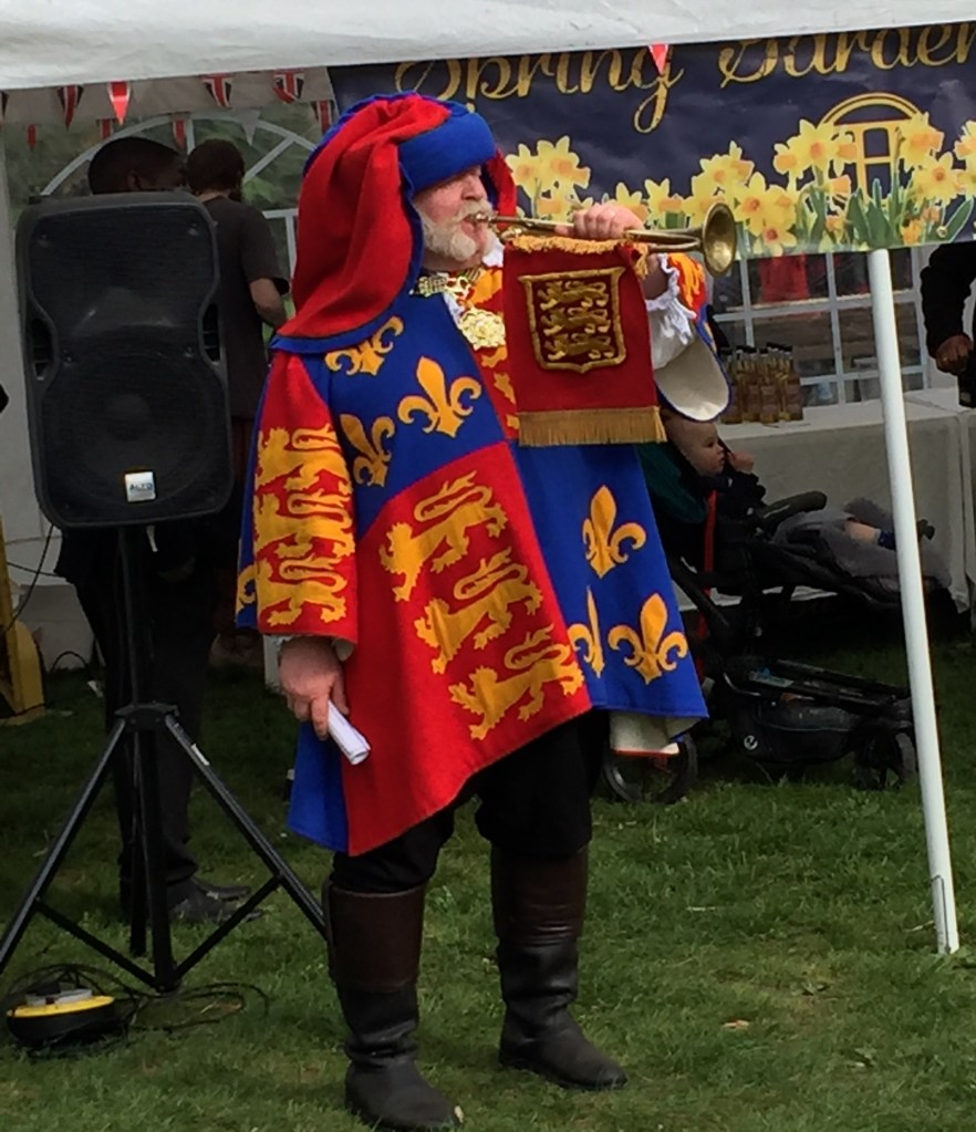 An announcer wearing a jacket designed like the Royal Standard flag, with colours of red, blue and yellow. He's blowing a trumpet, which has a red flag with yellow decorations hanging from it.