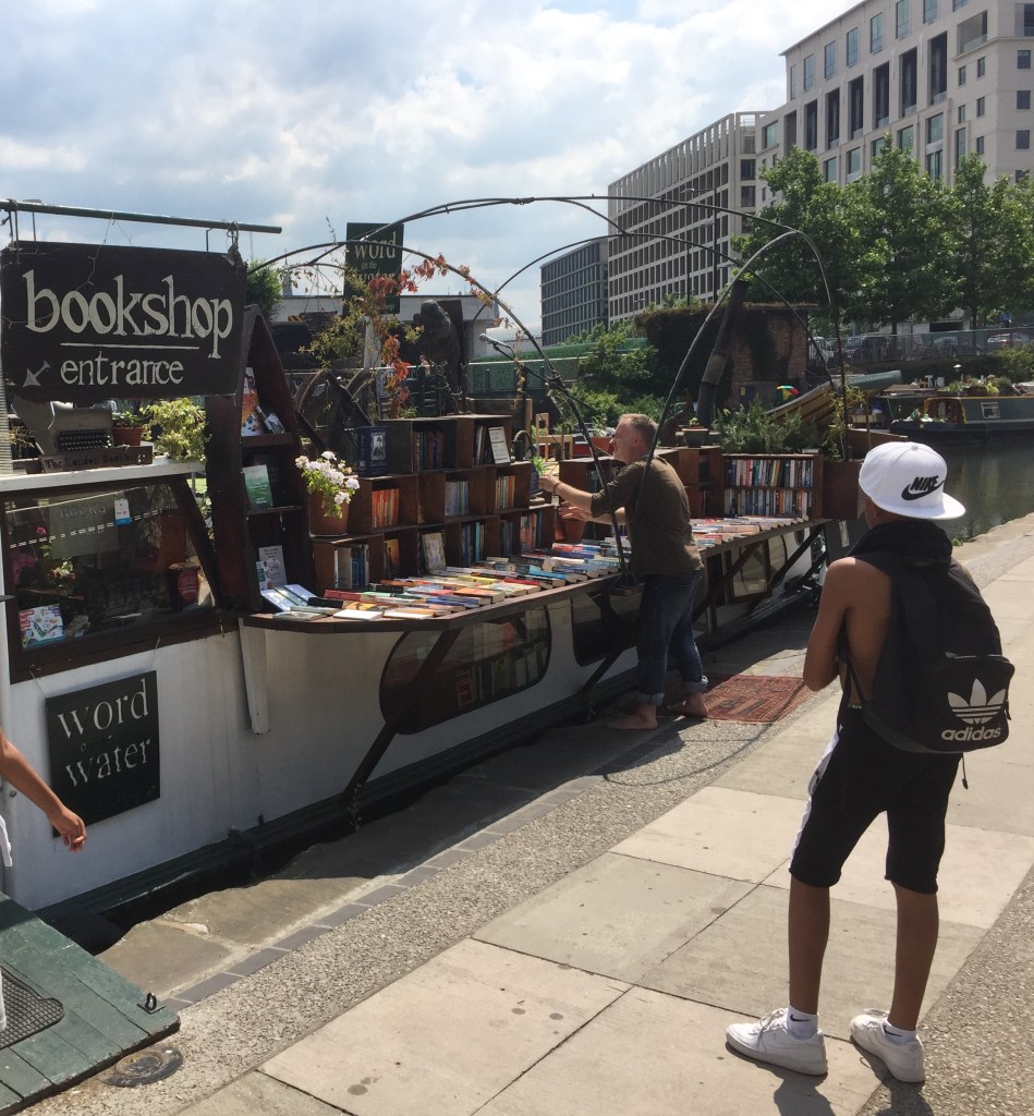 A bookshop on a canal boat on the King's Cross Canal, with shelving on the side of the boat showing hundreds of titles for sale.