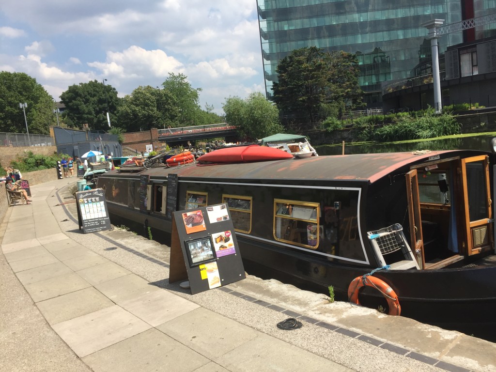 A canal boat on the King's Cross Canal, with chalkboard notices on the side of the vessel and A-board signs on the pathway next to it advertising a variety of cakes.