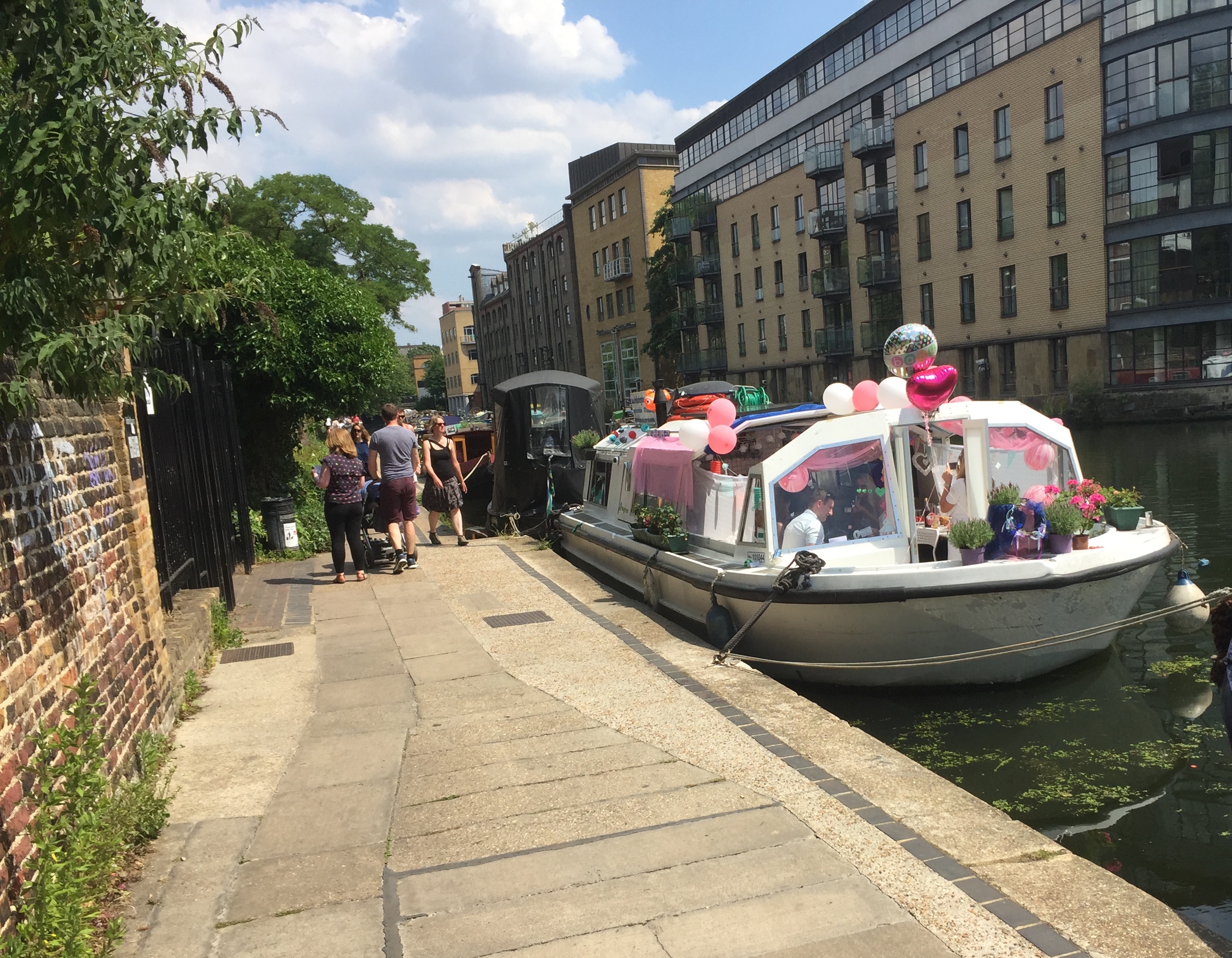 A white boat on the King's Cross Canal, with pink and white balloons attached to it. One balloon is in the shape of a heart, and there's also a polka dot balloon with the word baby on it.