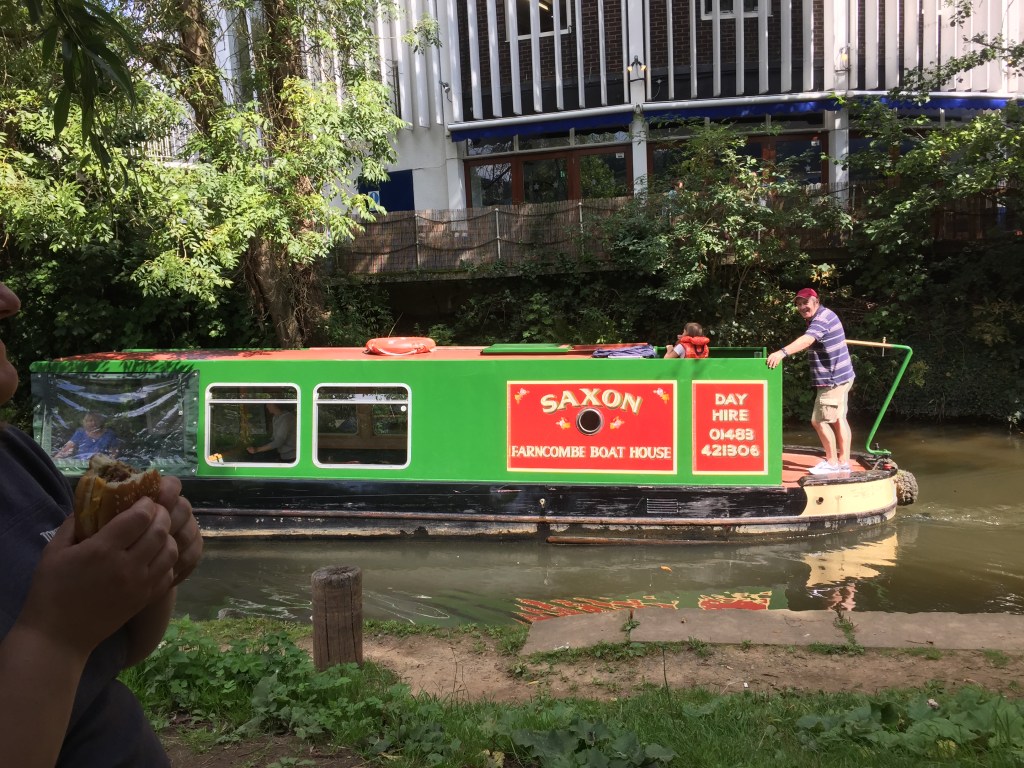 A man smiling over at us as he stands on the end of a long canal boat sailing down the River Wey in Guildford. The body of the boat is green, but has red signs with white lettering that read Saxon, Farncombe Boat House, Day Hire, 01483 421306.