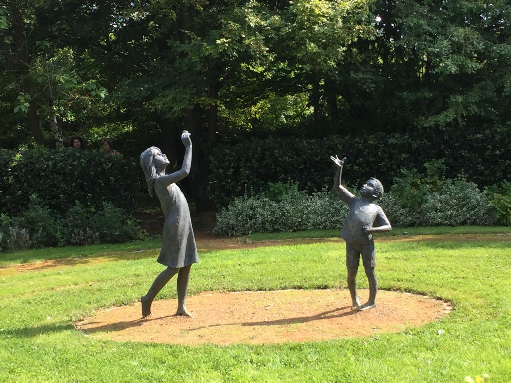 Black statues of a girl and boy playing on the grass outside Guildford Cathedral. They each have a hand raised, as if the girl is about to throw a small object for the boy to catch.