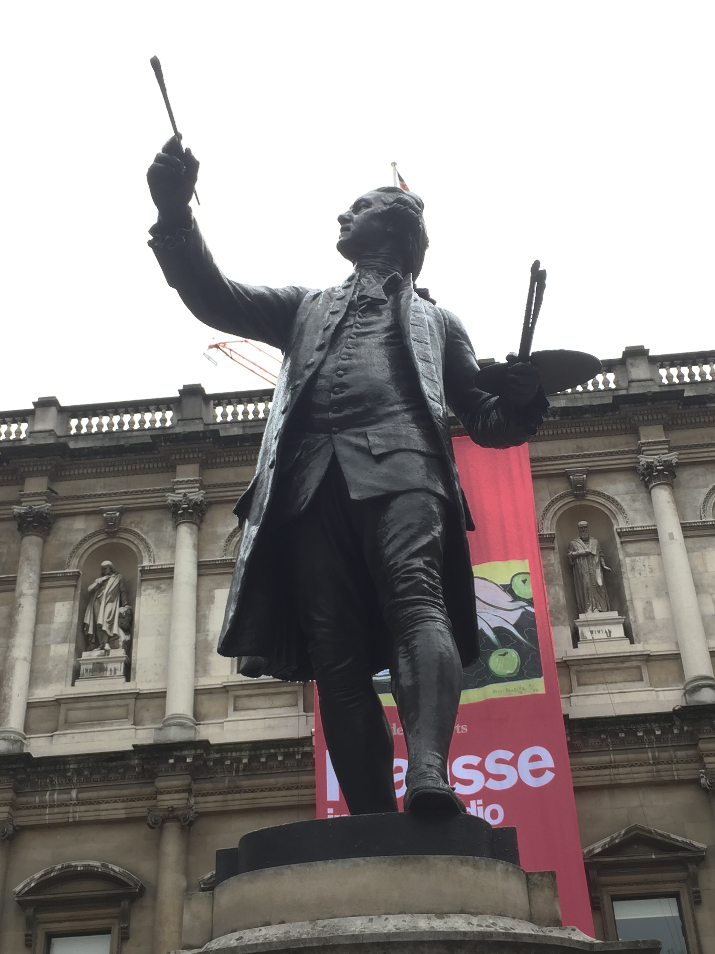 Statue of Sir Joshua Reynolds outside The Royal Academy Of Arts. He is holding a palette with a couple of paintbrushes sticking out of it in his left hand, while looking up at a paintbrush he's holding up high with his right hand.