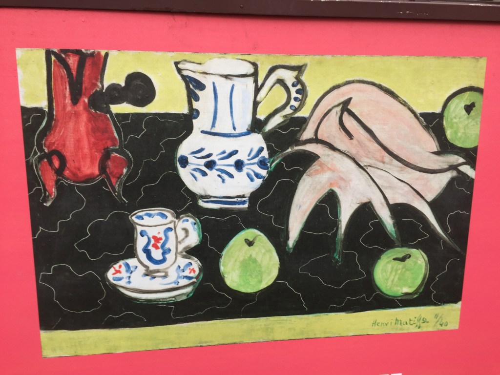Close-up of a collage artwork by Matisse, called Still Life With A Shell, on a poster for an exhibition about the artist at the Royal Academy Of Arts. The artwork includes a large grey representation of a seashell, as well as a white jug, mug and saucer that each have leafy blue detailing, plus a couple of green apples.