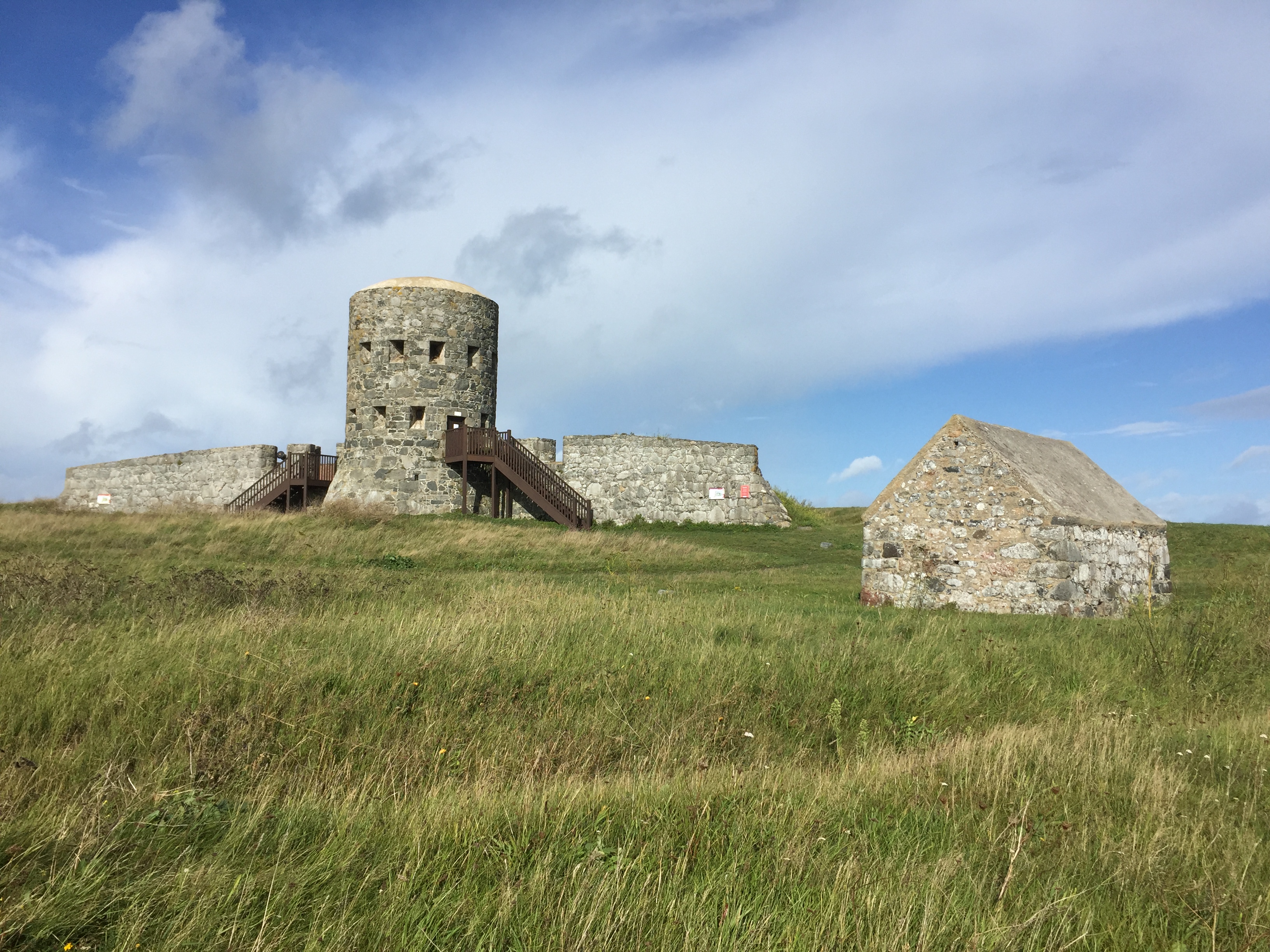A round stone tower in Guernsey, with small windows and a flight of wooden steps leading up to a doorway. The tower is flanked by low stall walls on each side, and there's a small house made of similar stonework nearby.