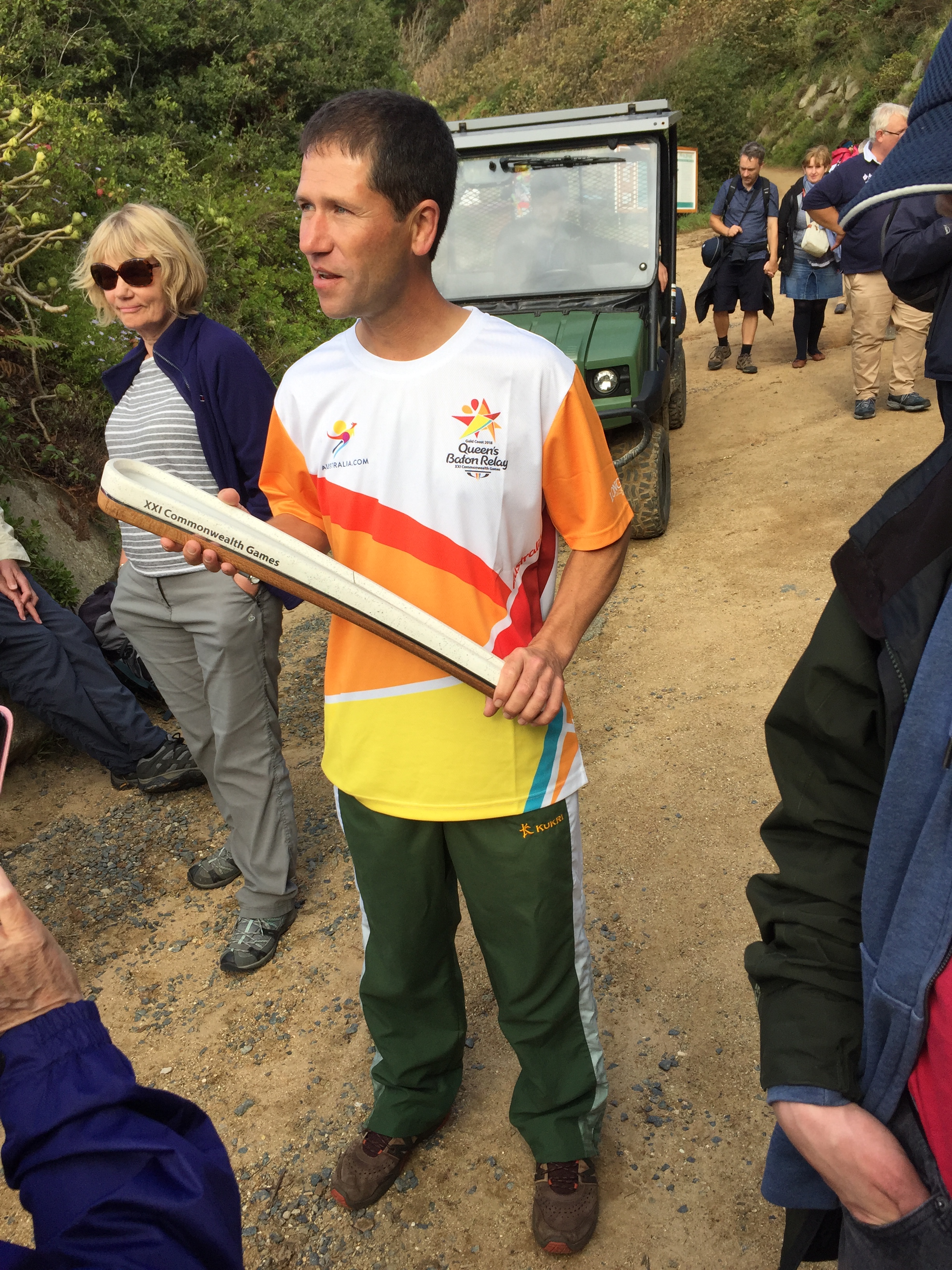 A man holding a wooden baton for the 21st Commonwealth Games. The baton is made of a layer of white wood on top of a layer of darker wood, and the overall shape of the baton is like an oversized and elongated eye of a needle, starting from a point at one end to a narrow curved opening at the top. Small black text on the light side of the baton says XXI Commonwealth Games. The man is wearing a t-shirt with white, red, orange and yellow colours on it, with a small colourful logo on each breast. On the right is a star-shaped logo, with black text that says Gold Coast 2018, Queen's Baton Relay, XXI Commonwealth Games. The other logo is a stylised version of an athlete running, and has Australia.com beneath it.