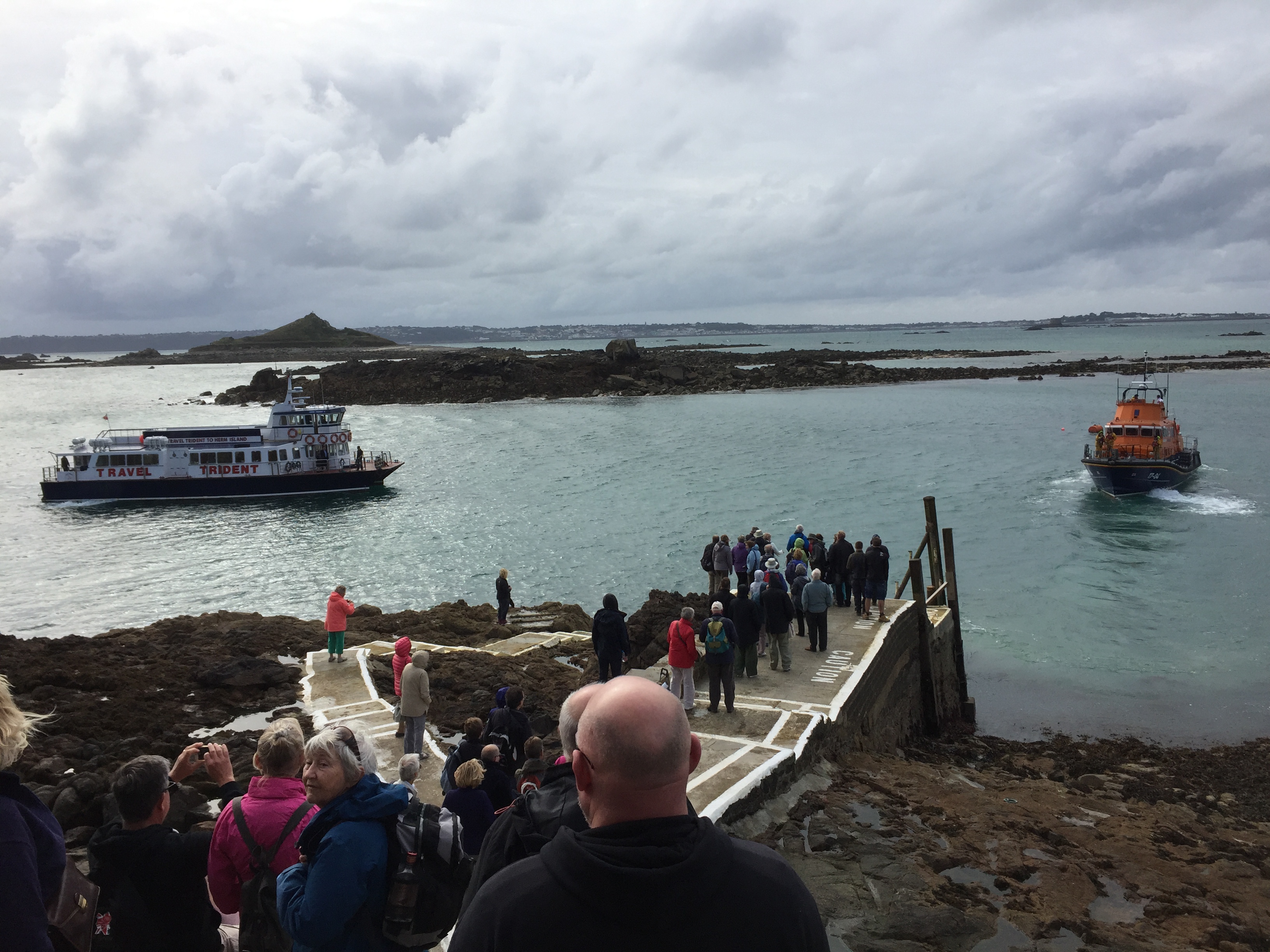 People waiting on a stone flight of steps on Herm Island for a small ferry approaching that the words Travel Trident. An orange lifeboat ferry is also nearby. Small rocky outcrops can be seen in the background, and the island of Guernsey on the horizon.