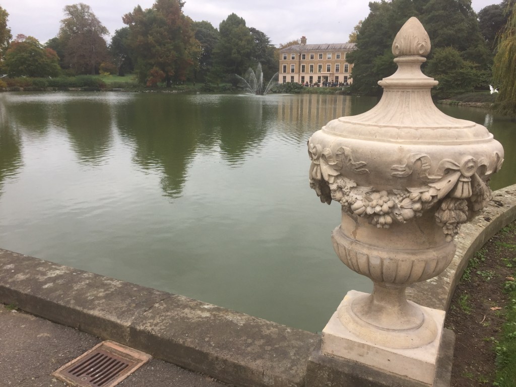 A large ornate stone urn overlooking the wide lake at Kew Gardens, in which there is a fountain near the far shore.