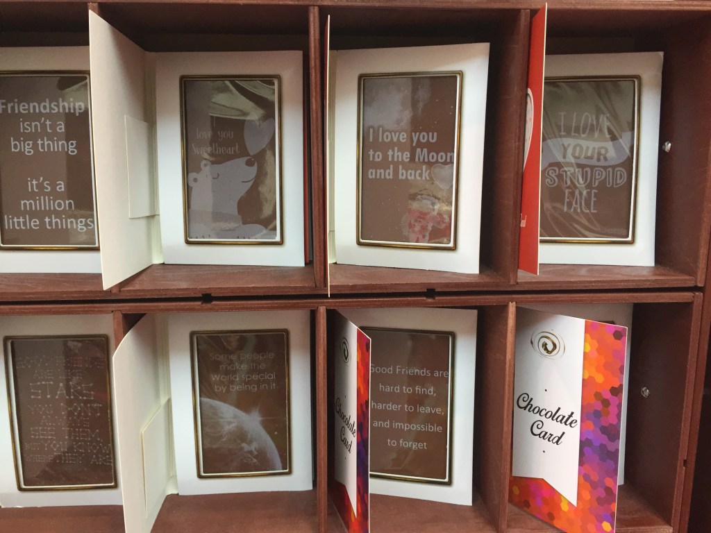Shelves of open cards where the message page has a plastic window, behind which is a large slab of chocolate with a message in white on it. For example, friendship isn't a big thing, it's a million little things. I love you sweetheart. I love you to the moon and back. I love your stupid face. Some people make the world special by being in it. Good friends are hard to find, harder to leave, and impossible to forget.