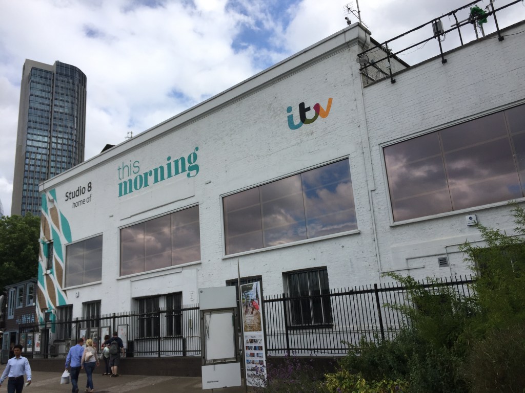 A long 2-storey white building with wide windows along its length. Wording above the windows reads Studio 8, home of This Morning, along with the ITV logo, which has the letters in lowercase with rainbow colours across them.