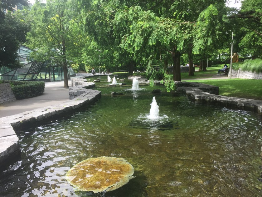A long curving body of water in Jubilee Park in Canary Wharf, enclosed by a low rock wall, containing a few small fountains along its length. It has trees hanging low overhead and there is a pathway running alongside, with more trees, grass and pathways visible in the distance.