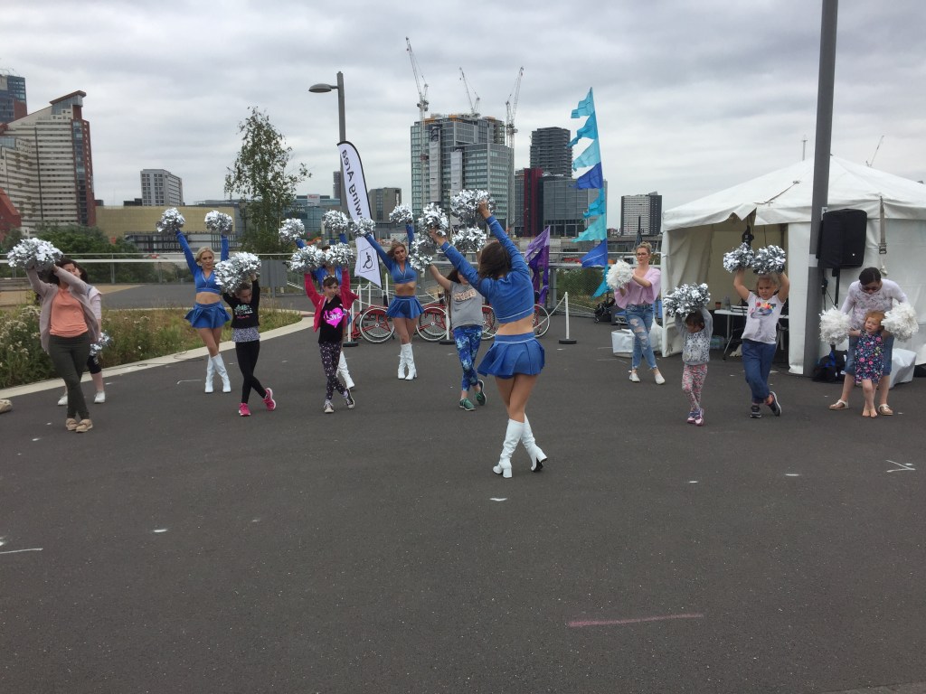 Cheerleaders performing a routine, wearing blue tops, short blue skirts and white boots, and waving silver pom-poms. A few members of the public, including adults and children, also have silver pom-poms as they join in.