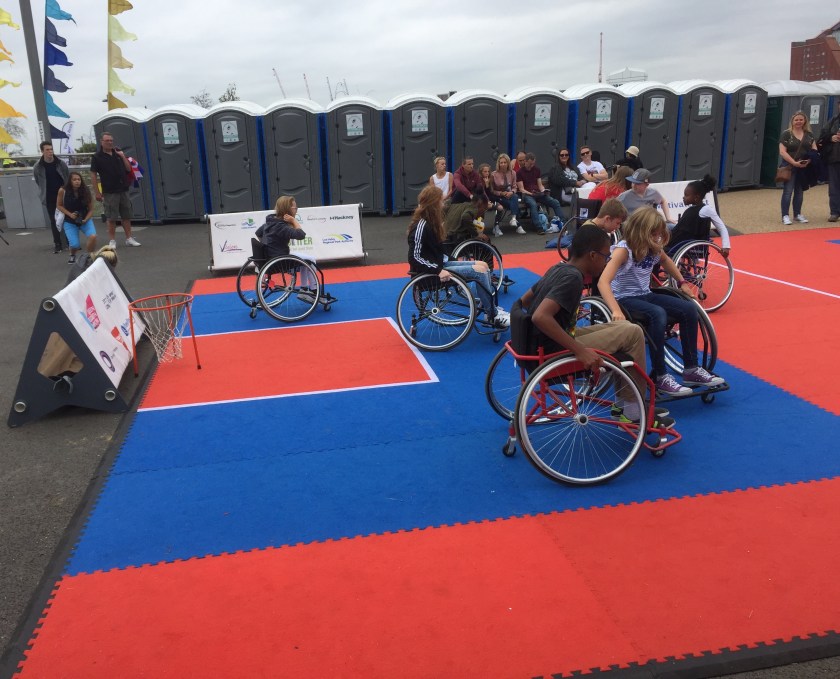 A group of people playing wheelchair basketball in the Olympic Park, on a pitch divided into red and blue sections, with the basket at floor level.